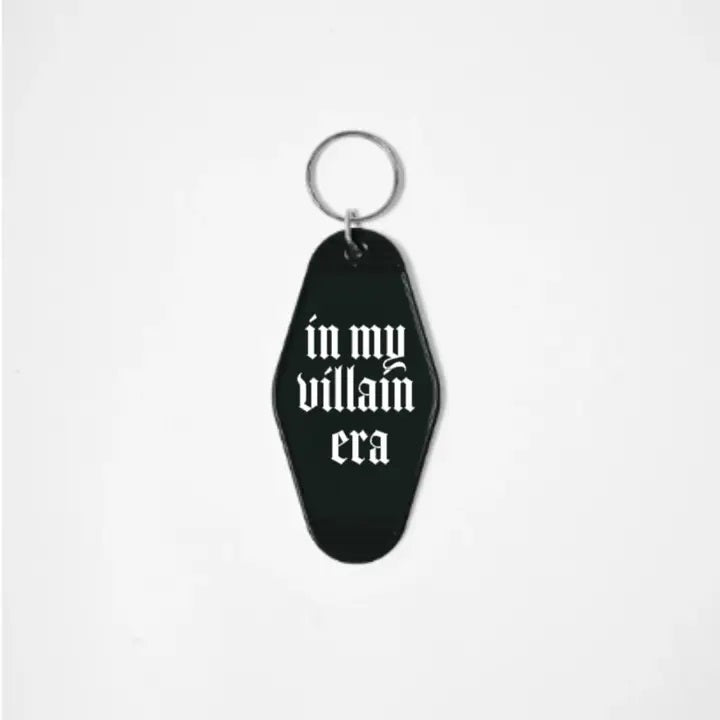 A black keychain shaped like a motel tag with the words "In My Villain era" in a white gothic font