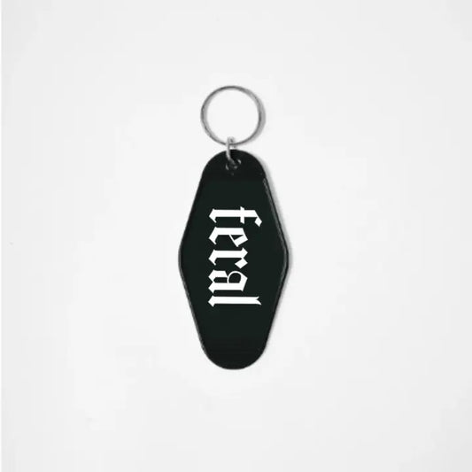 A black keychain shaped like a motel tag with the word "Feral" written in a white gothic font