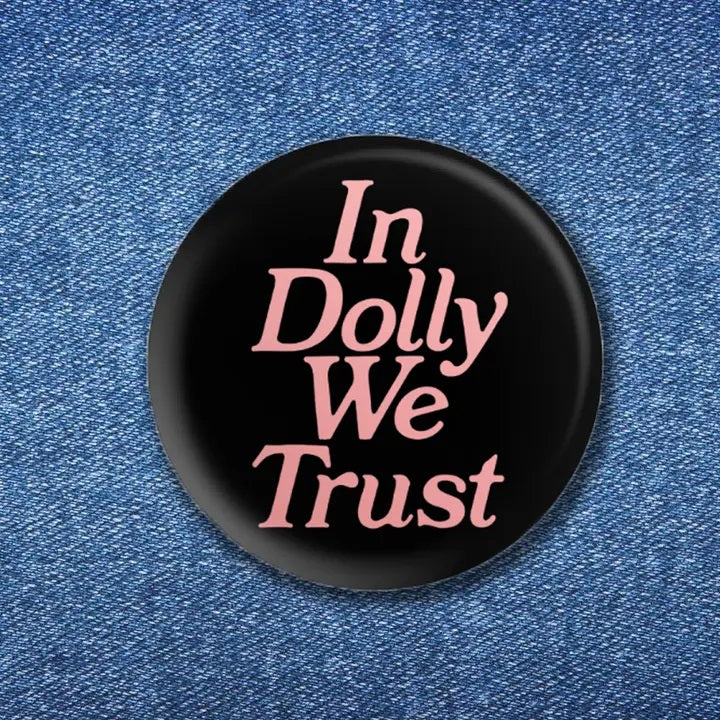 A black button pin with the words "In Dolly We Trust" written in a pink font