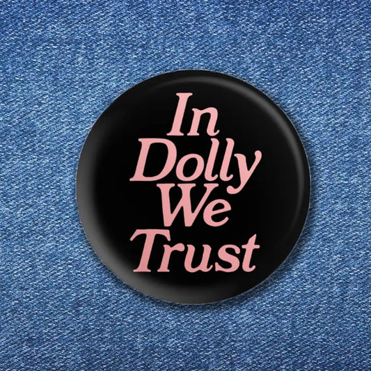 A black button pin with the words "In Dolly We Trust" written in a pink font