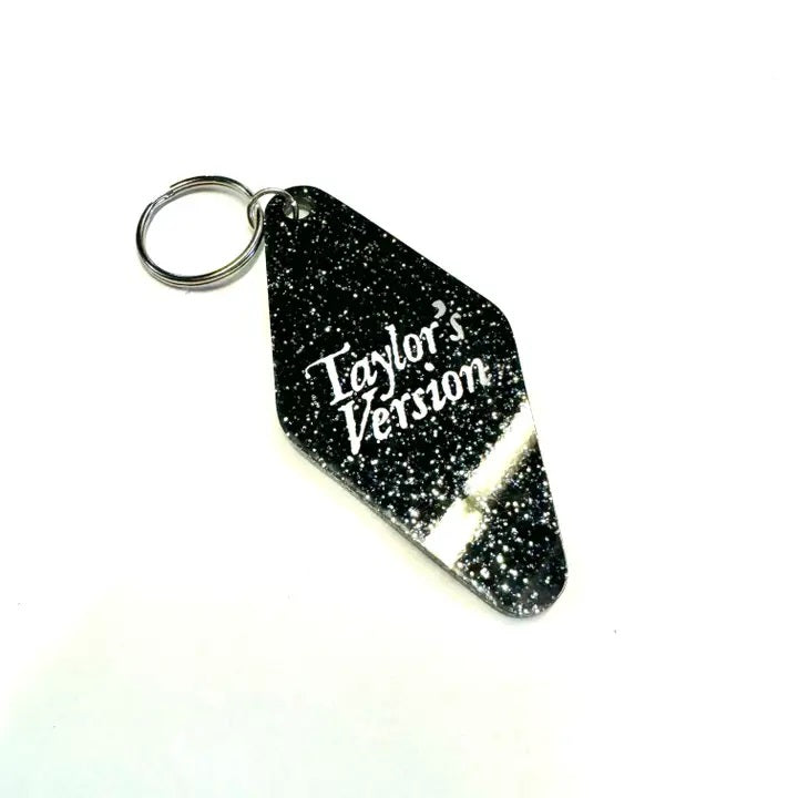 A black glitter keychain shaped like a motel tag with the words "Taylor's Version" in white font