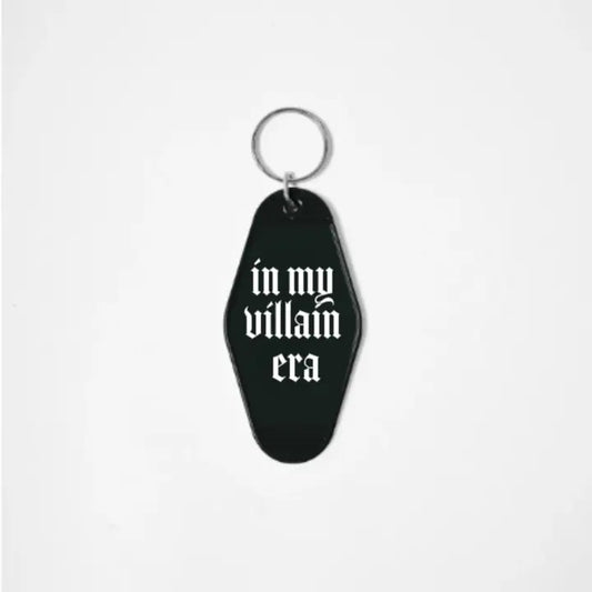 A black keychain shaped like a motel tag with the words "In My Villain era" in a white gothic font